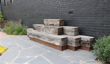A stack of 6 large timber beams of various lengths raised off the ground on top of a plinth and resting against a brick wall serving a dual purpose, as a visual feature part of the landscaping and also a rest spot to sit