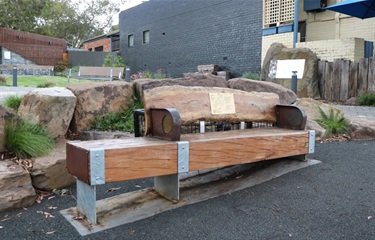 Crafted street furniture bench seat made from timber and steel, with the seat base resembling a large timber beam paired with a seat back that has an organic finish, featuring the natural knots and intricacies of the source tree that was repurposed.